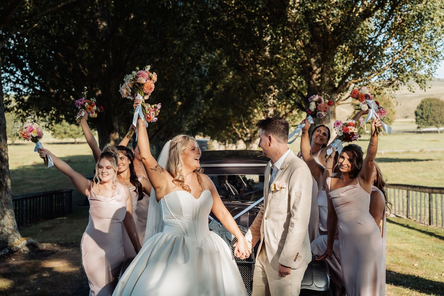 Bride and groom celebrate with bridesmaids raising colorful bouquets beside a vintage black car outdoors.