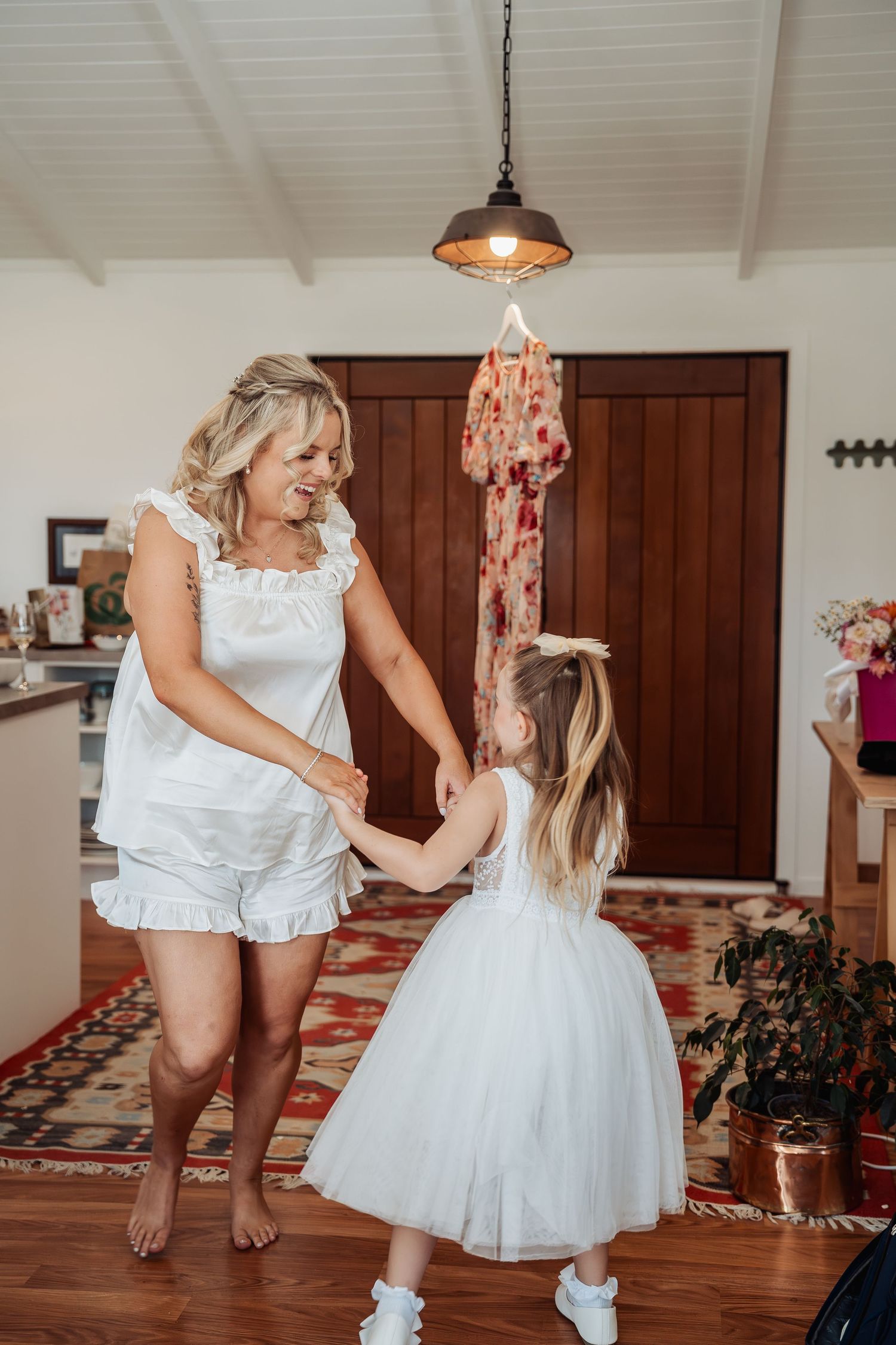 A woman in white dances with a young flower girl in a tulle dress inside a warmly decorated home.