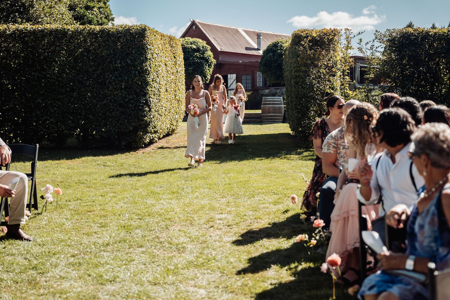 Bridesmaids in blush dresses walking down a garden aisle lined with wedding guests.