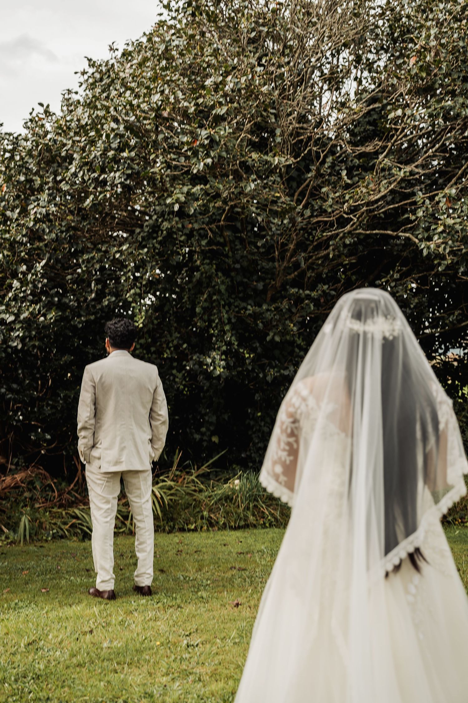 Bride in lace wedding dress and veil approaches groom in beige suit during outdoor first look ceremony.