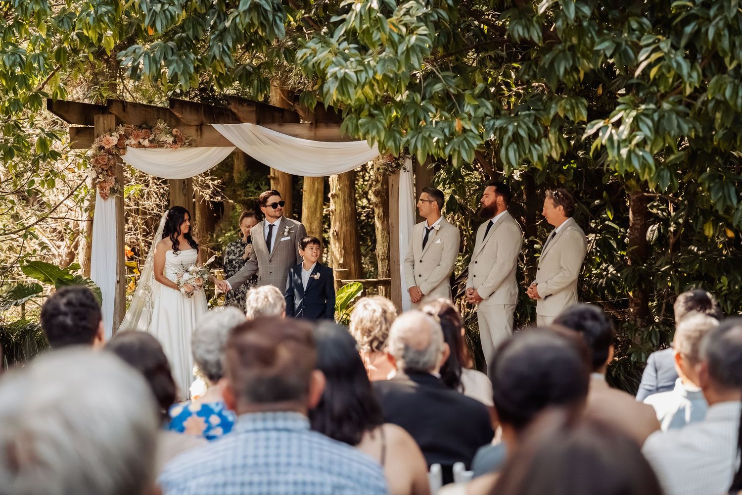 Wedding guests watch as couple exchanges vows beneath a rustic floral arbor in a sunlit garden.