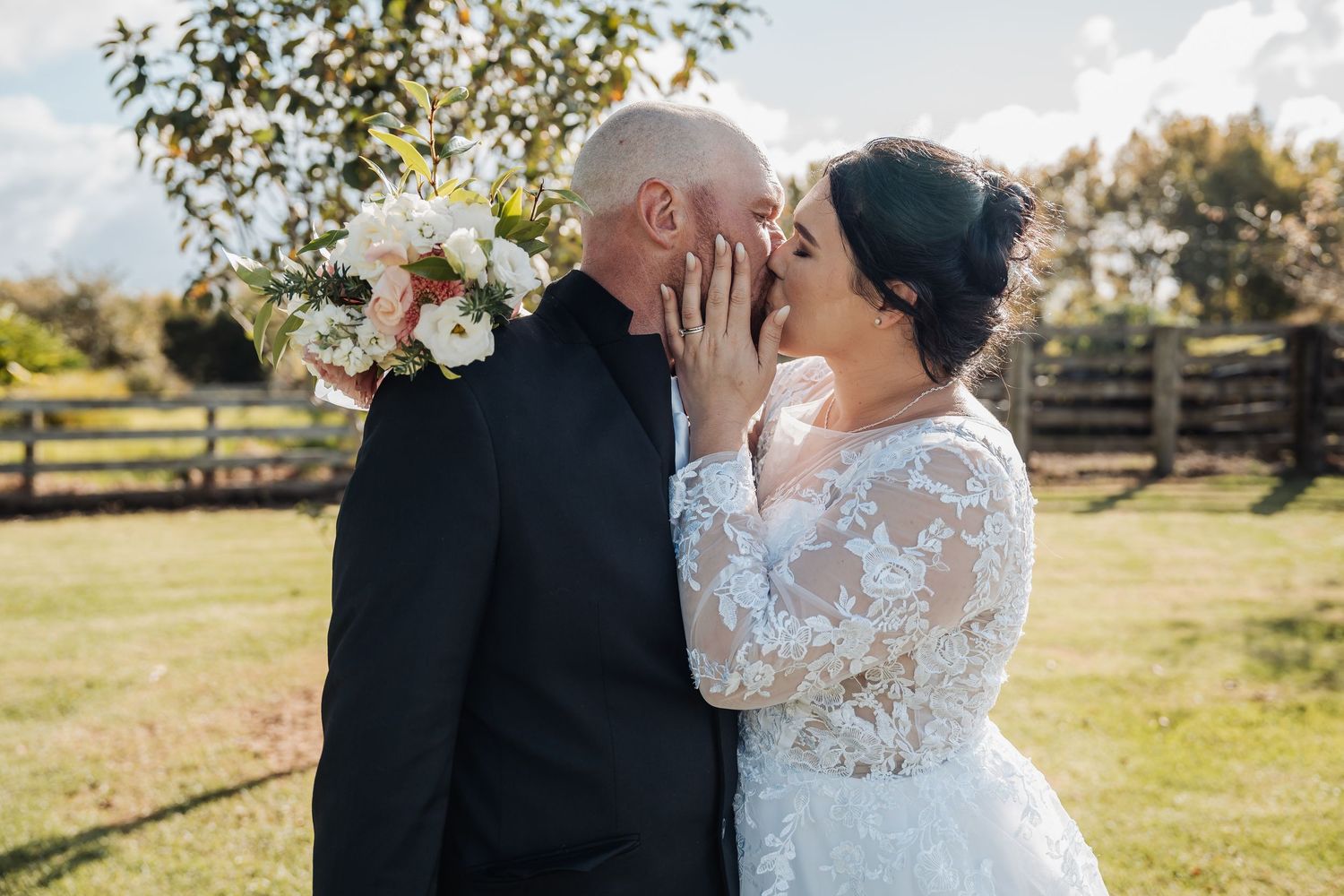 Tender wedding moment captured in sunny outdoor setting with rustic wooden fence backdrop.