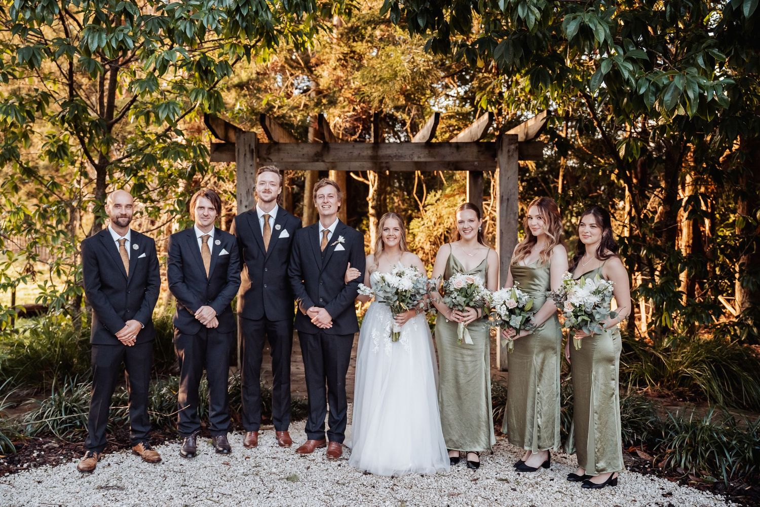Wedding party poses outdoors under wooden arch with bridesmaids in sage dresses and groomsmen in navy suits.