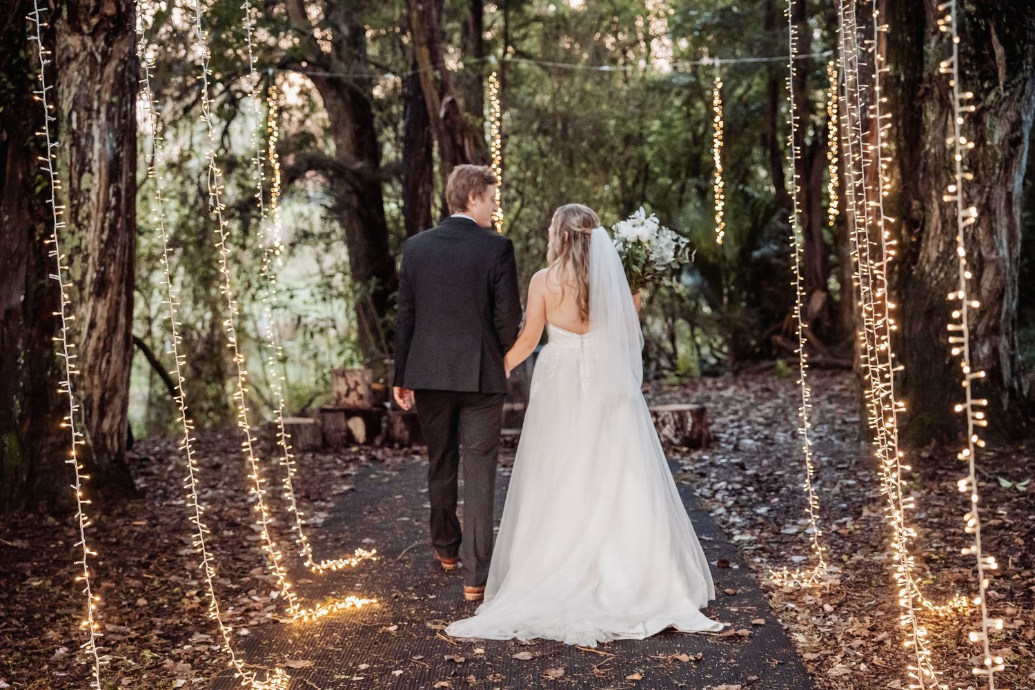 A newlywed couple walks down an enchanted forest path illuminated by twinkling string lights.