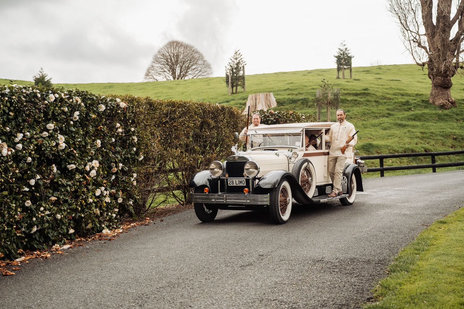 Vintage cars line a curved driveway at a countryside wedding venue with green hills in the background.
