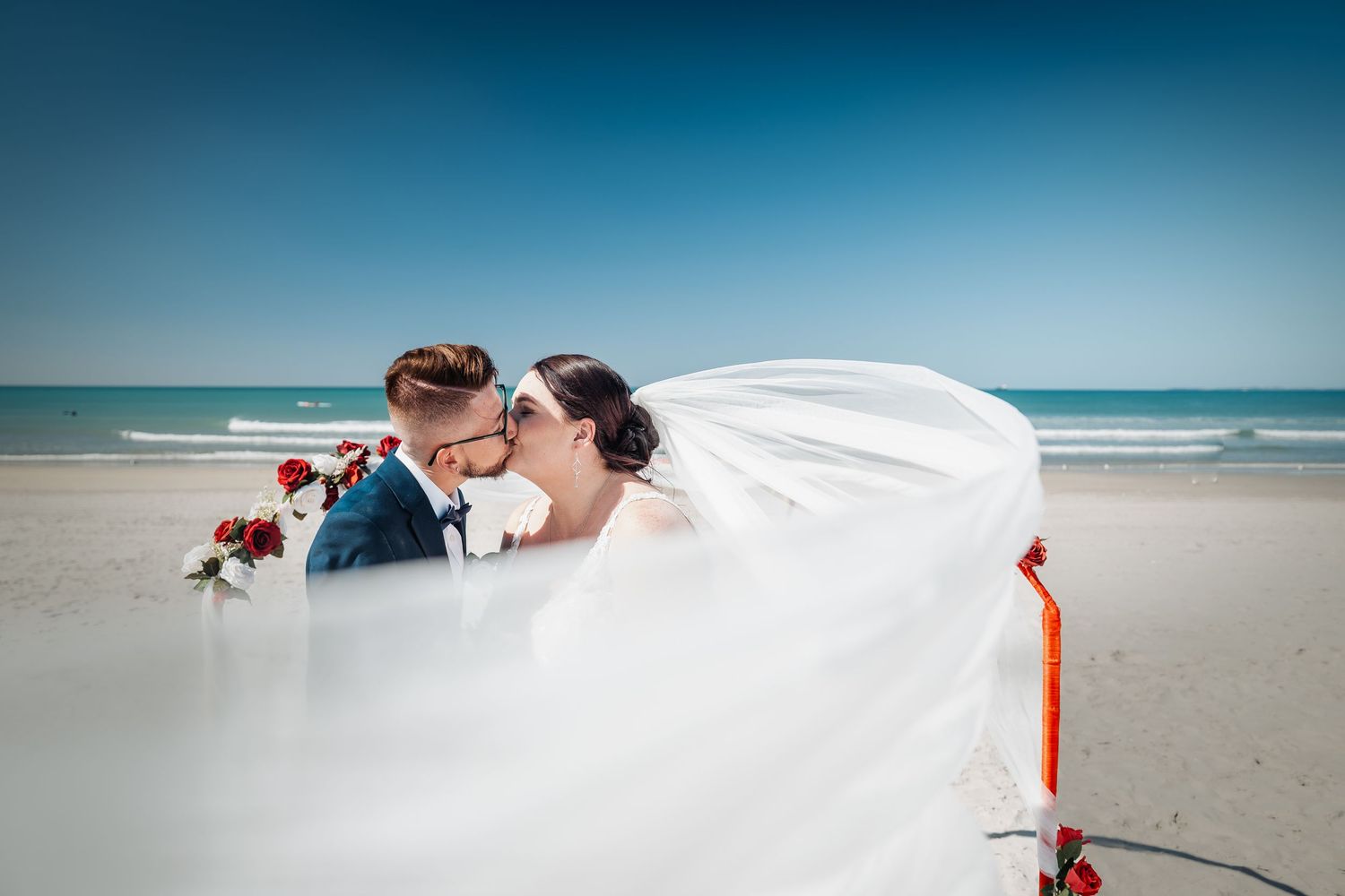 A romantic wedding couple embraces on a beach as a flowing white veil catches the ocean breeze.