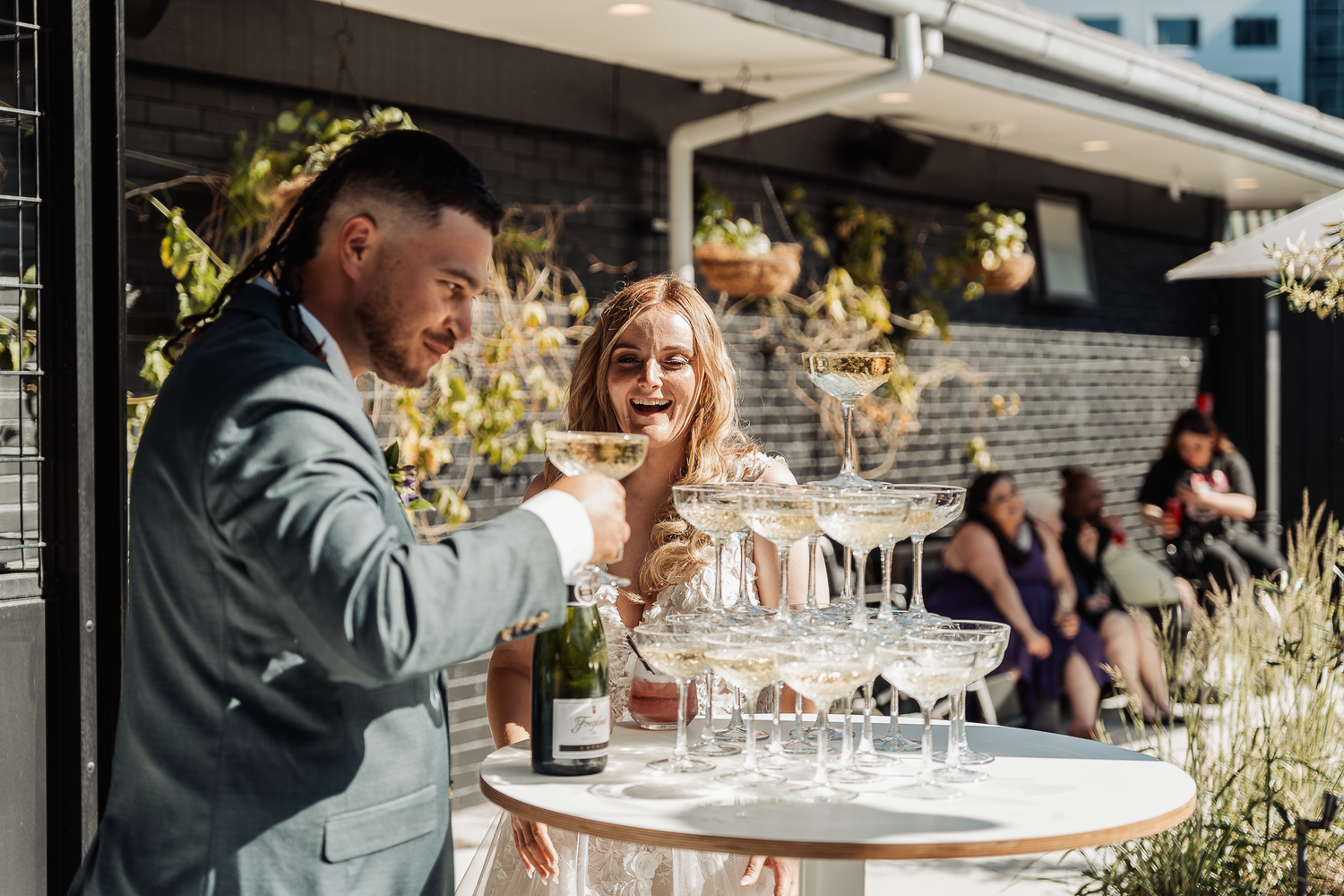 Newlyweds share champagne at an outdoor reception with a tower of glasses on a white table.