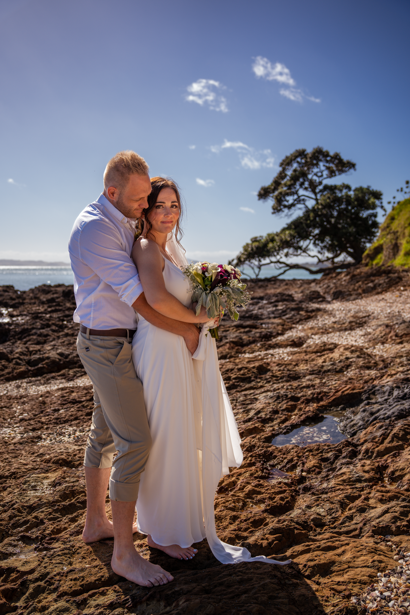 A romantic seaside wedding photo with a couple embracing on rocky coastal terrain.