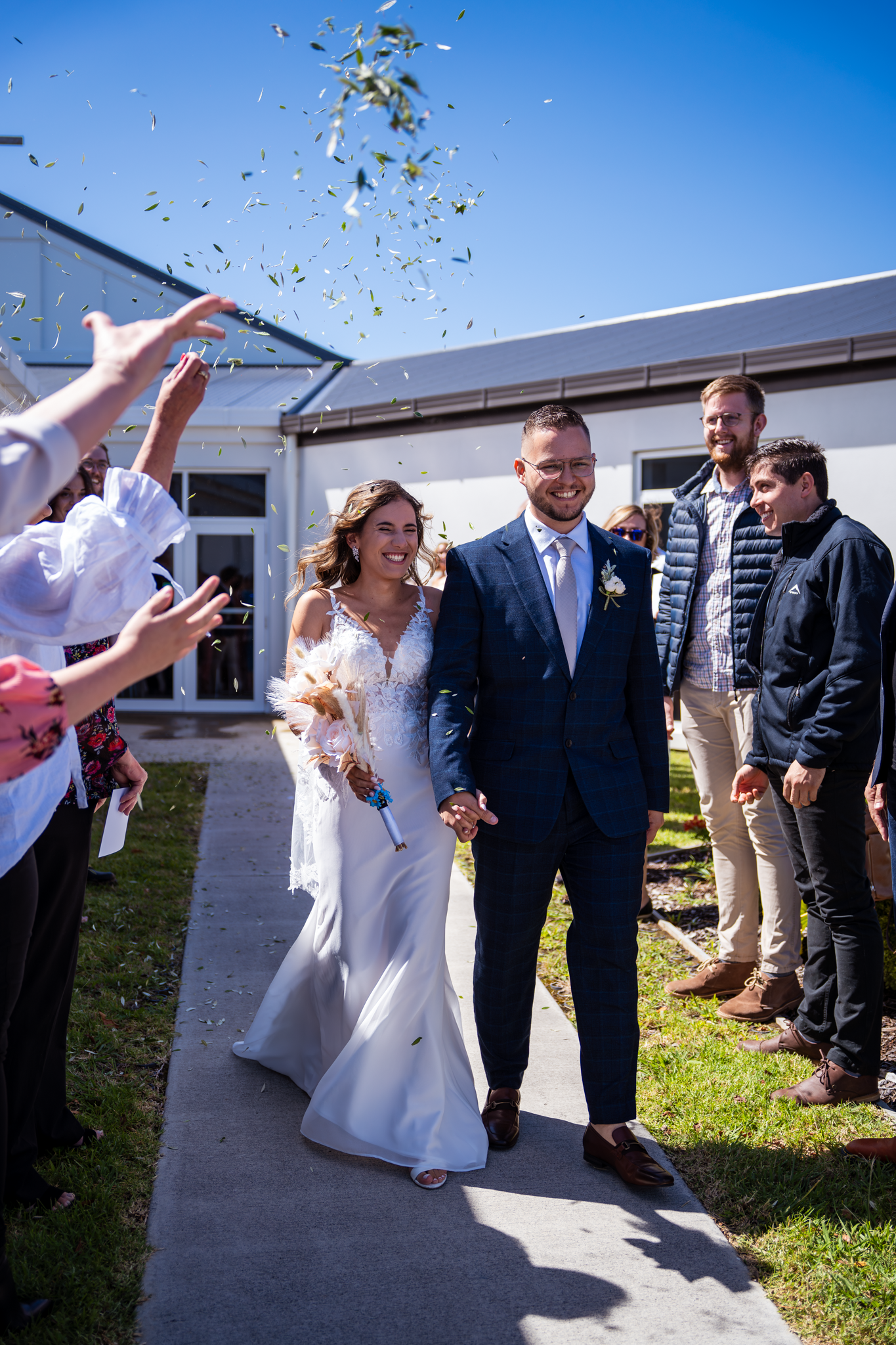 A joyful wedding exit with guests throwing confetti as a couple walks down an outdoor pathway on a sunny day.