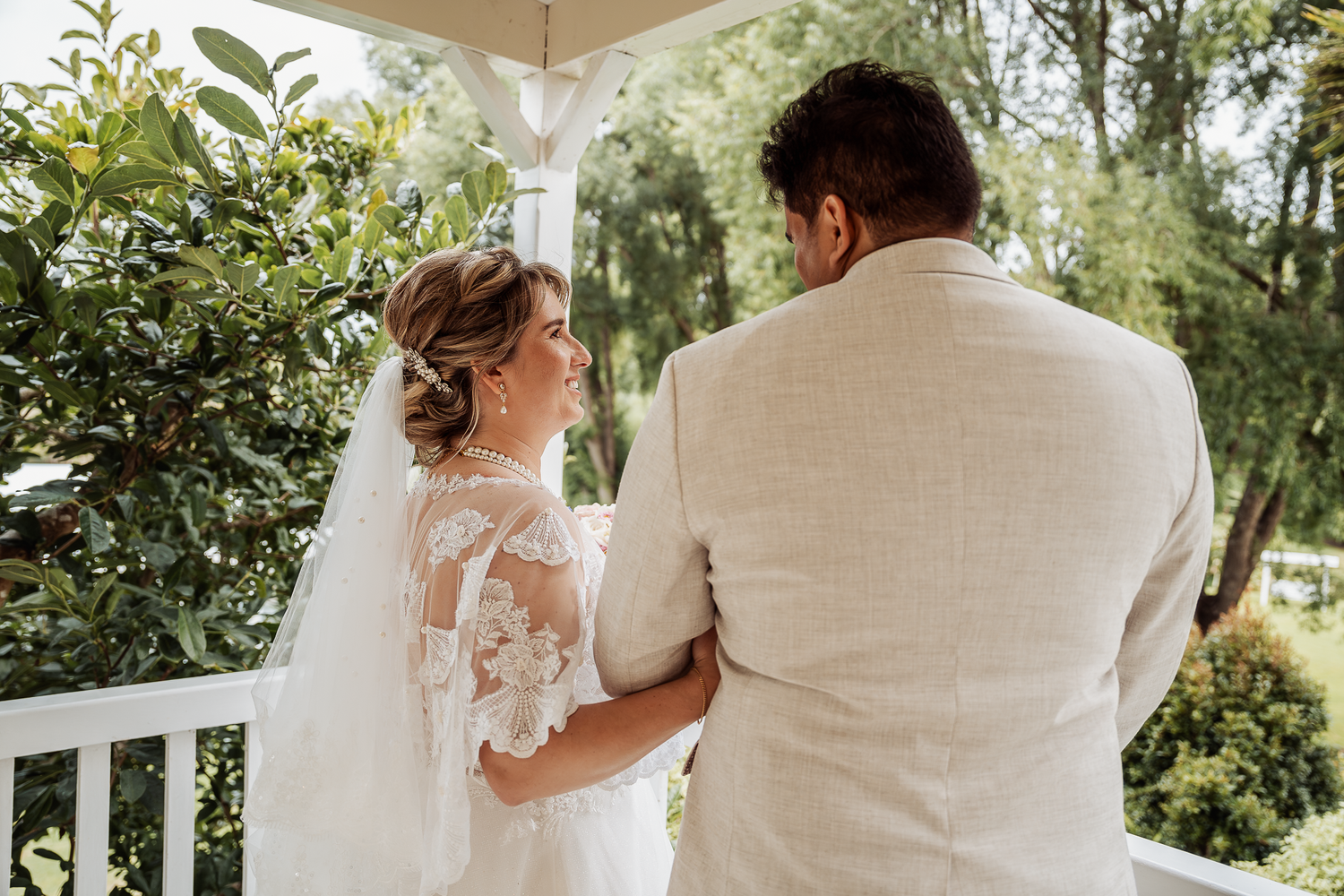 Couple in wedding attire stands together on white porch surrounded by greenery.