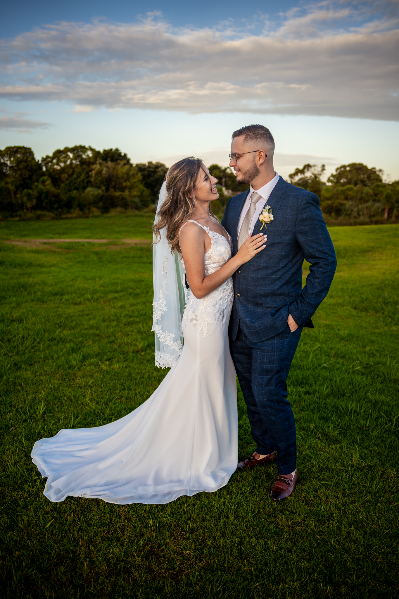 A romantic wedding portrait of a couple in formal attire on a grassy field at sunset.