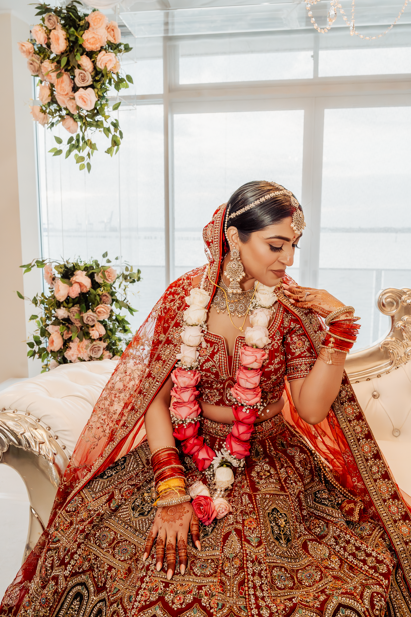 Bride in ornate red lehenga poses on elegant white sofa with floral decor.