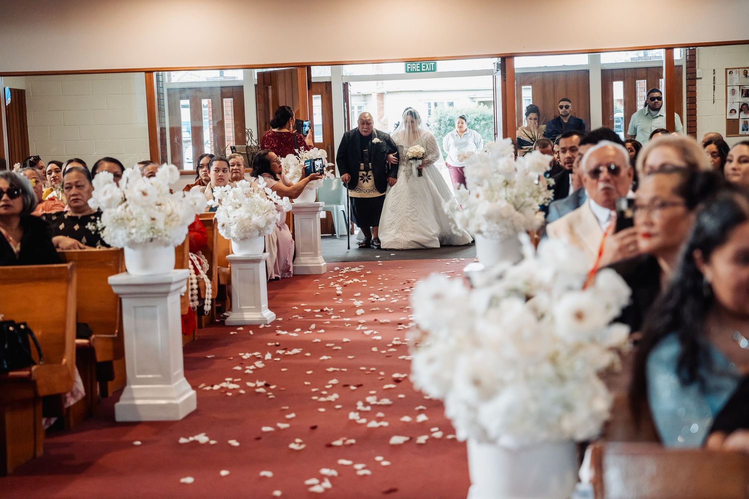 Wedding ceremony guests seated in church pews with white flowers decorating the aisle.
