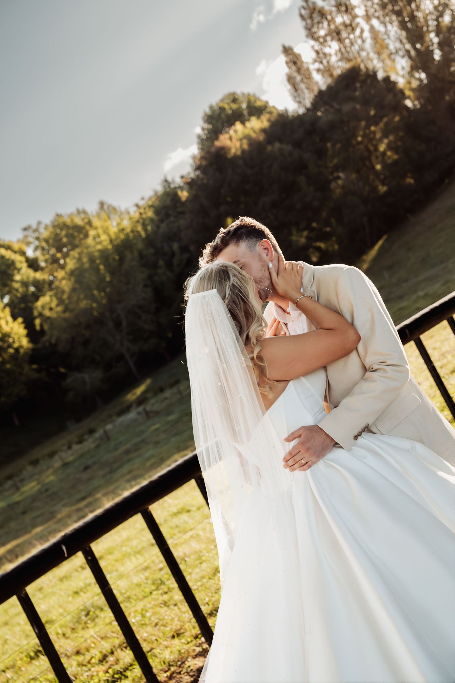 Bride and groom share a romantic kiss while embracing near a fence with lush green fields in the background.
