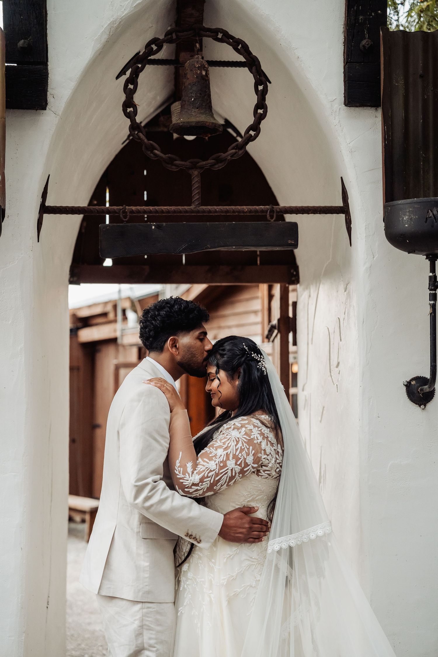Bride and groom share an intimate embrace under a rustic white arch with a vintage bell overhead.