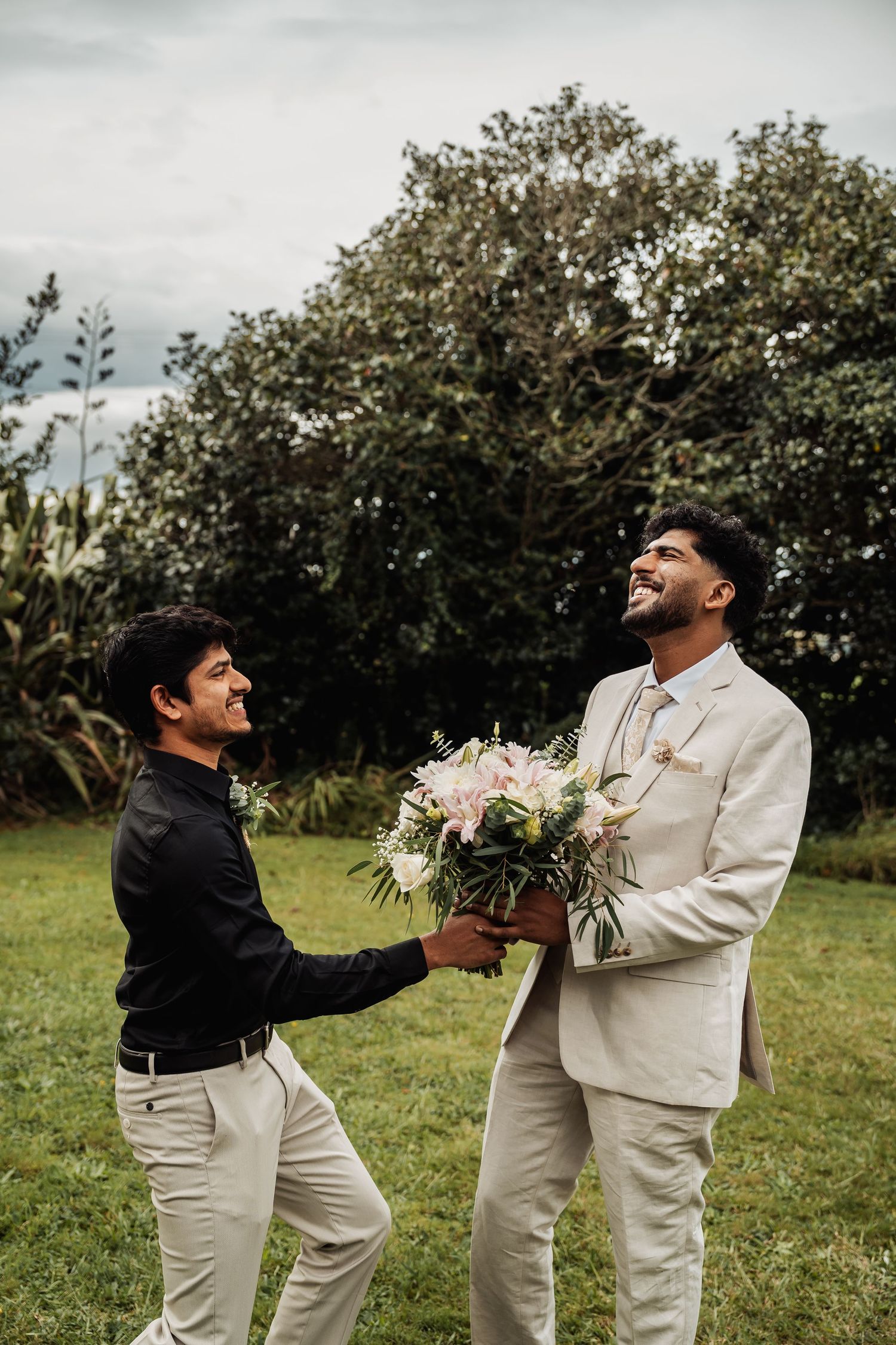 Two men laughing outdoors, groom in beige suit holding floral bouquet with groomsman.