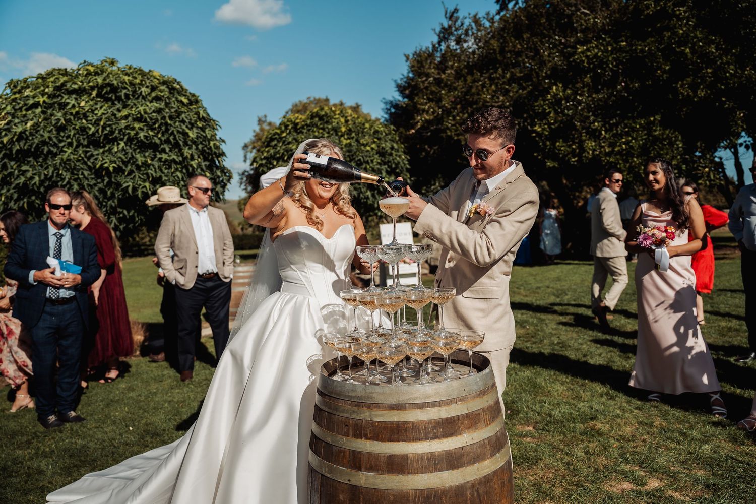 Bride and groom pouring champagne tower on barrel surrounded by wedding guests outdoors.