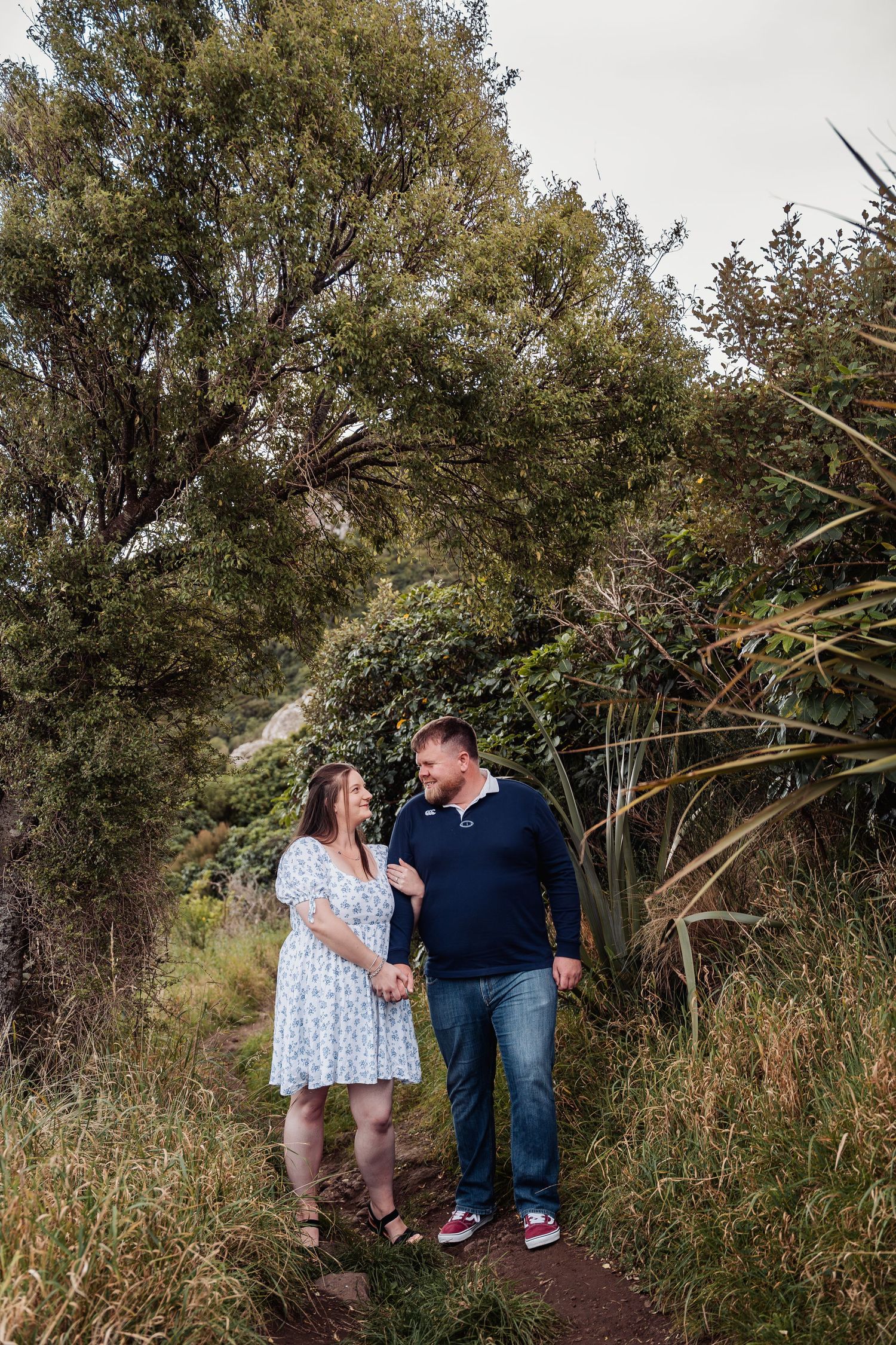 Couple holding hands and smiling at each other on a nature trail surrounded by lush green trees and plants.