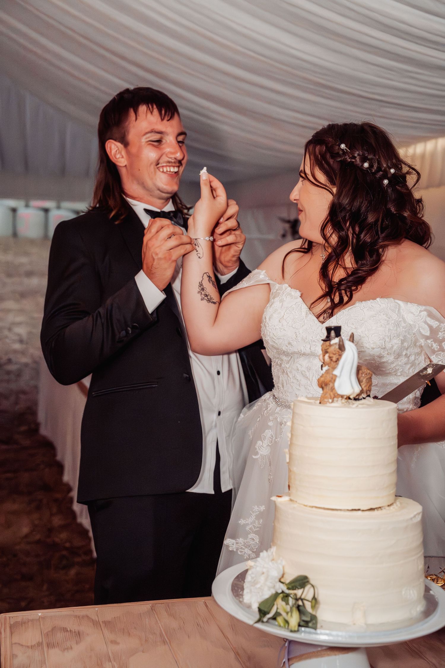 Laughing bride and groom feed each other cake at their wedding reception under a white tent.