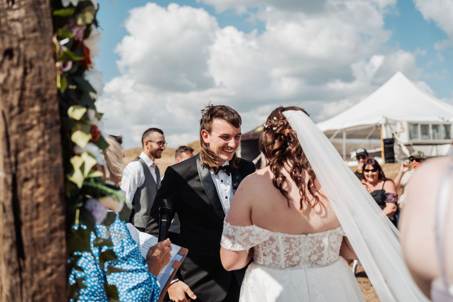 Groom smiling joyfully at bride during outdoor wedding ceremony with guests and white tent in background.