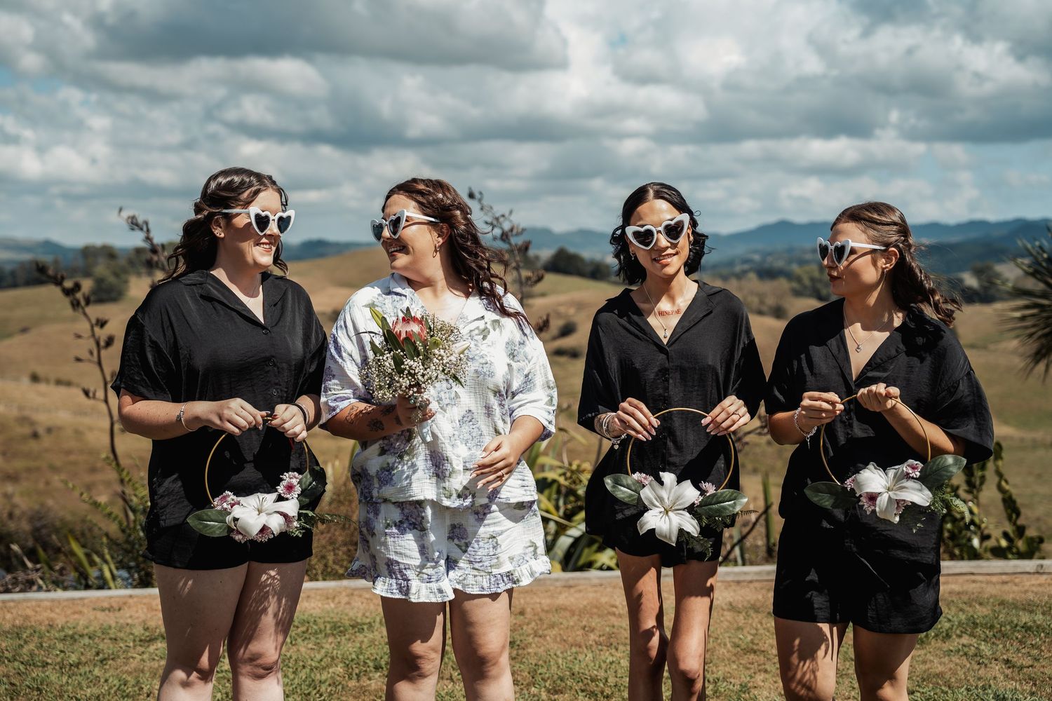 Four bridesmaids in black robes holding flowers and wearing sunglasses outdoors with scenic hills backdrop.