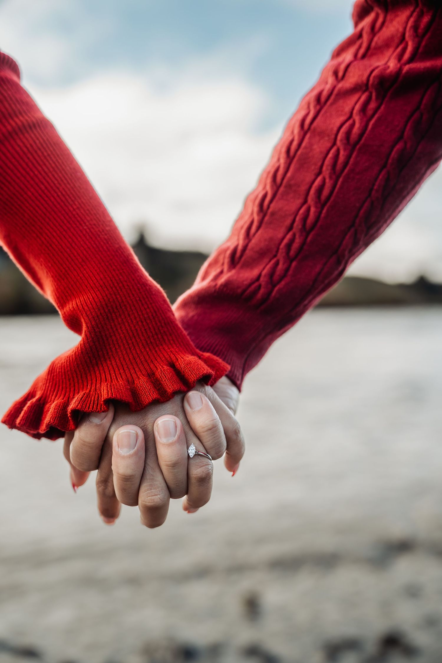 Couple holding hands wearing red sweaters with an engagement ring visible, standing near a beach on a cloudy day.