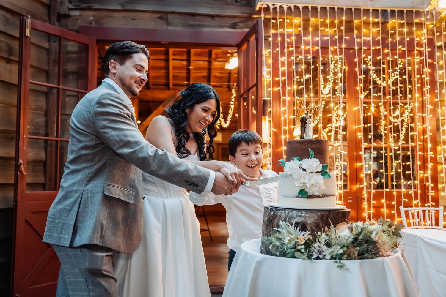 Bride, groom, and young boy cut wedding cake together in rustic barn venue with warm fairy light backdrop.