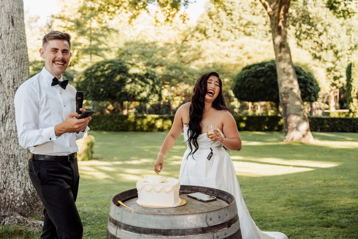 Laughing bride and groom cutting wedding cake on a rustic barrel outdoors in a garden setting.