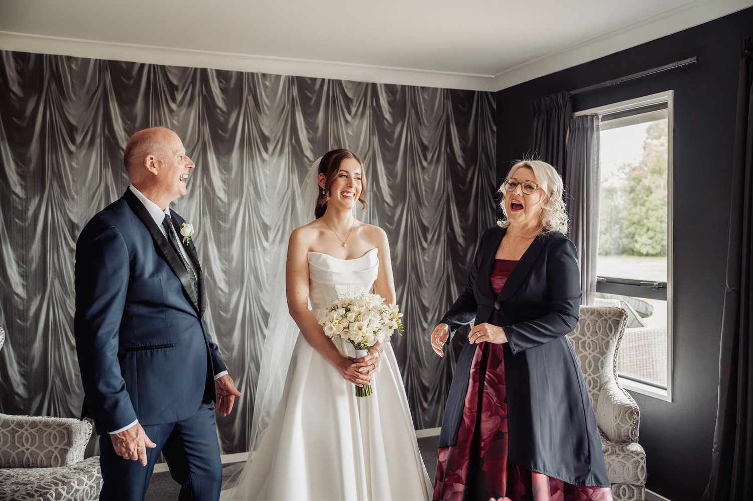 Bride in white gown holds bouquet, smiling with family in elegant grey-draped room during wedding preparations.