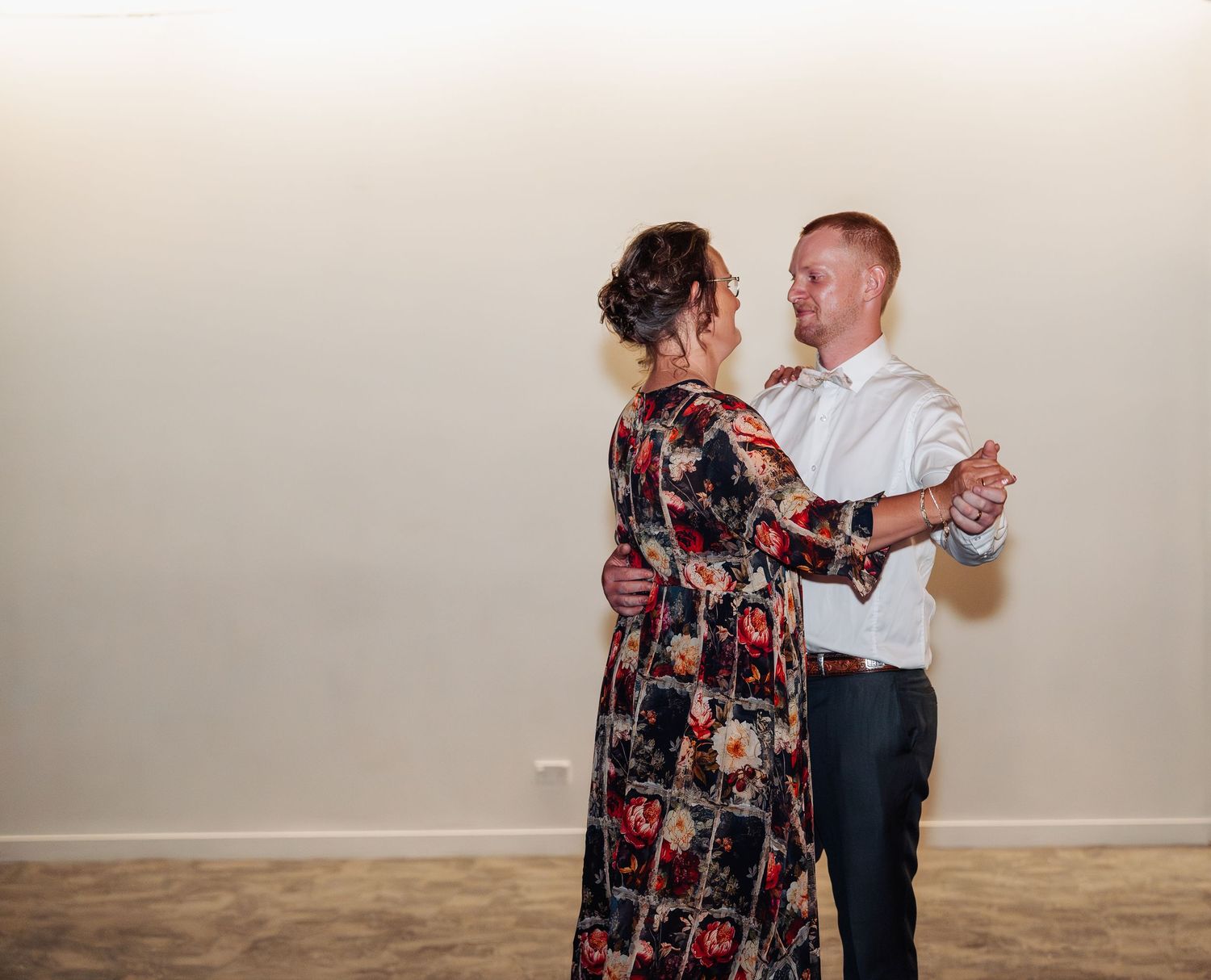 Couple shares a romantic dance together at a wedding reception, man in white shirt and woman in floral dress.