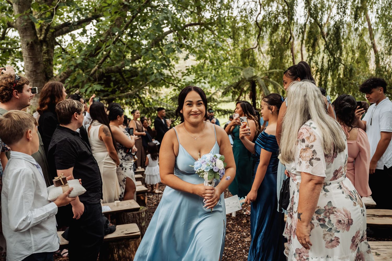 Bridesmaid in blue satin dress holding bouquet walks down outdoor wedding aisle surrounded by guests.