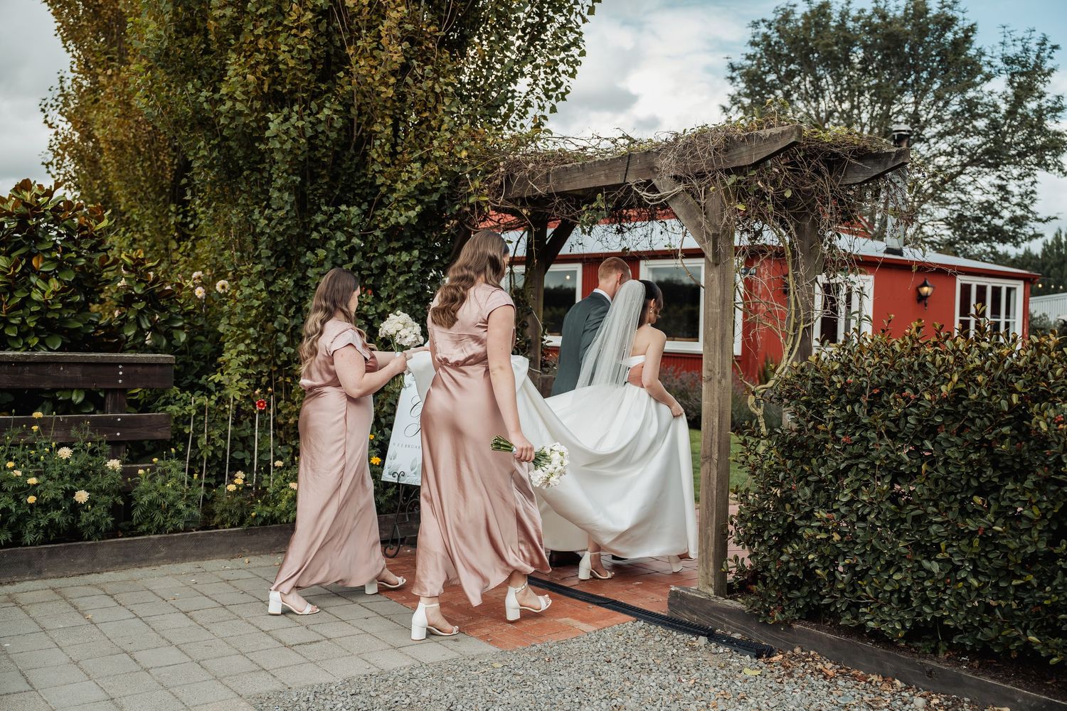 Bridesmaids in blush gowns helping bride with white dress in garden wedding venue.