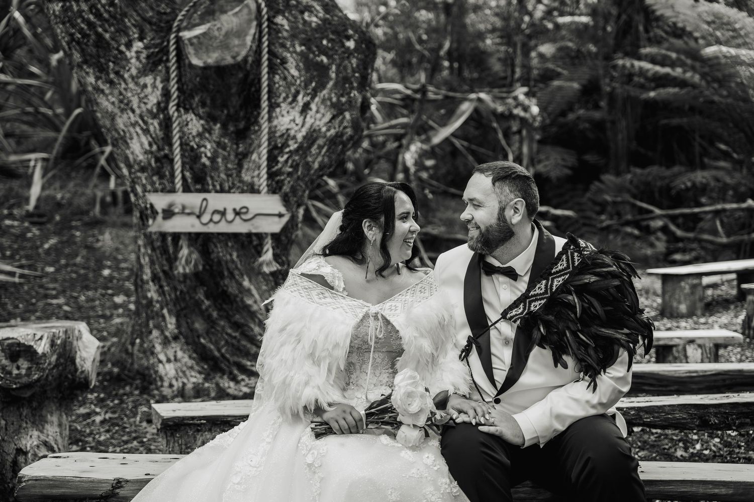 Black and white photo of bride and groom sharing a romantic moment on a bench in a forest setting.