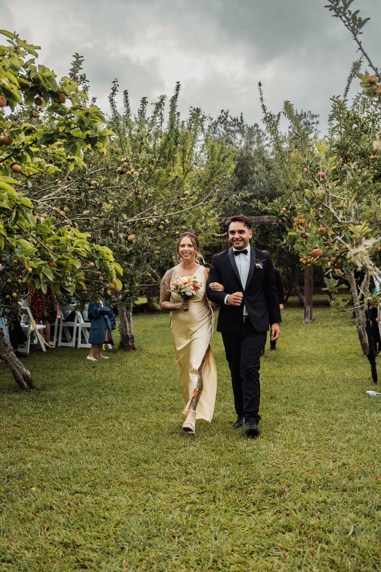 Bride and groom walk joyfully through an outdoor orchard wedding aisle surrounded by lush apple trees.