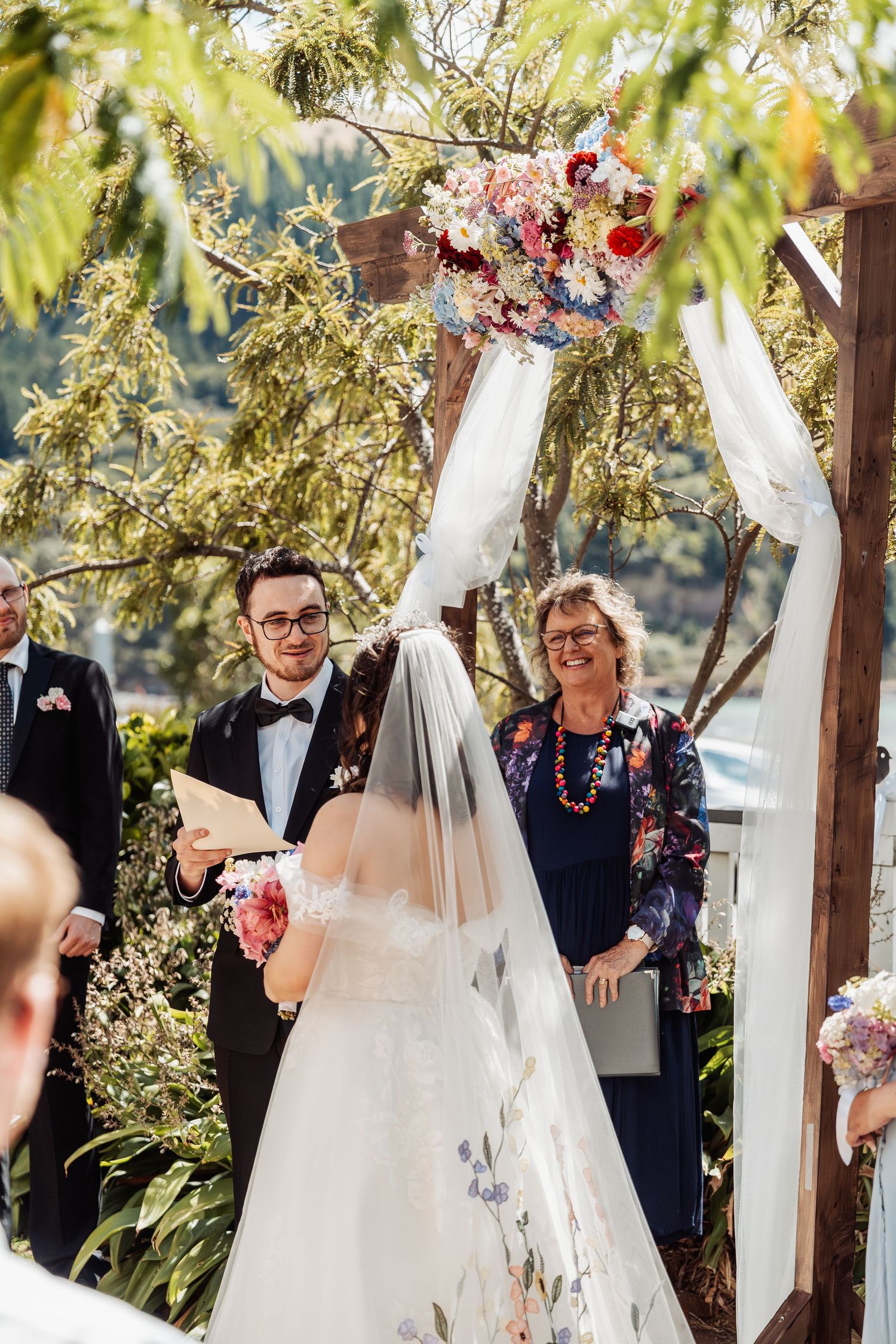 Groom reads vows to bride at outdoor wedding ceremony under floral arch with officiant smiling behind.