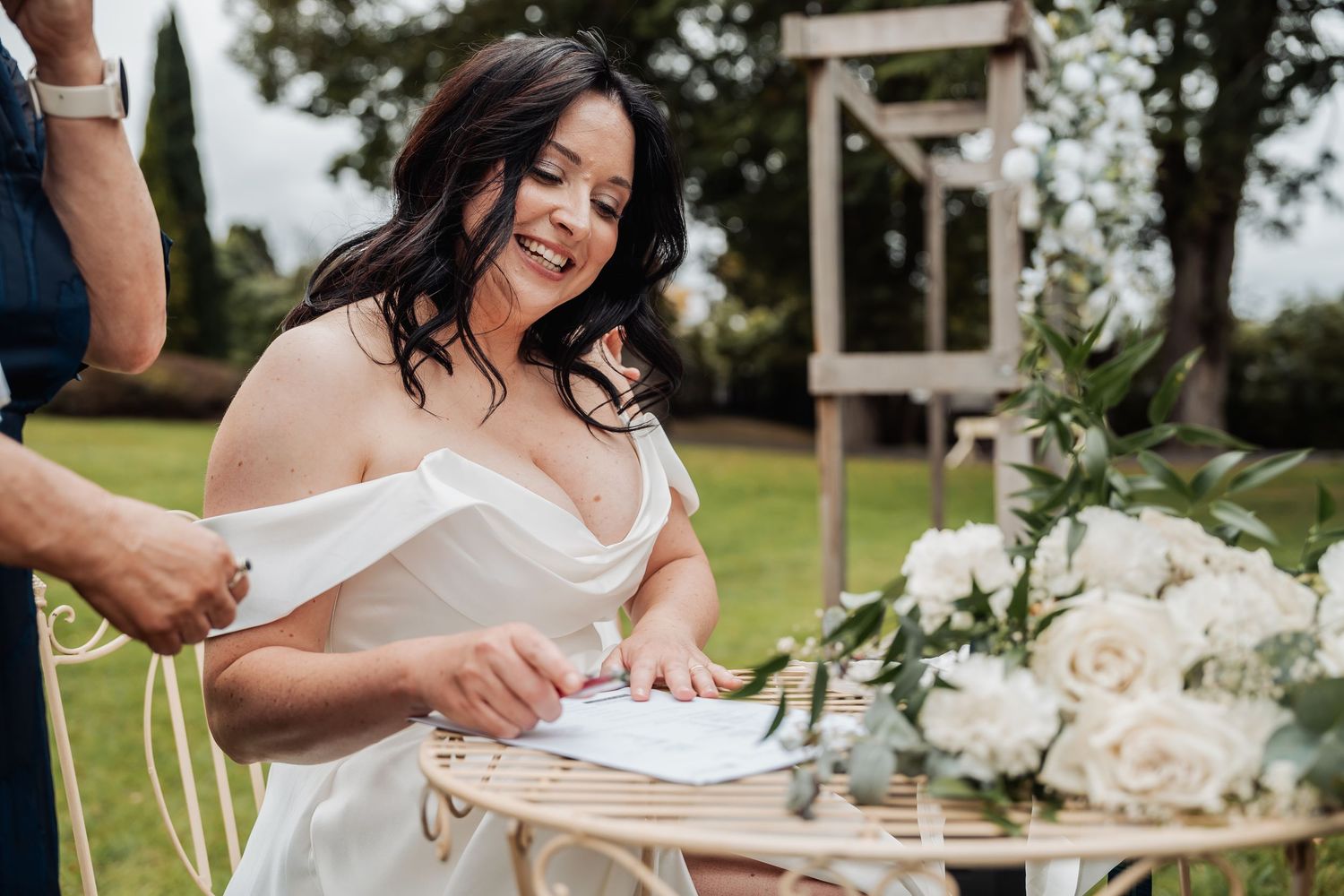 Smiling bride in white off-shoulder gown signing wedding documents at floral decorated table outdoors.