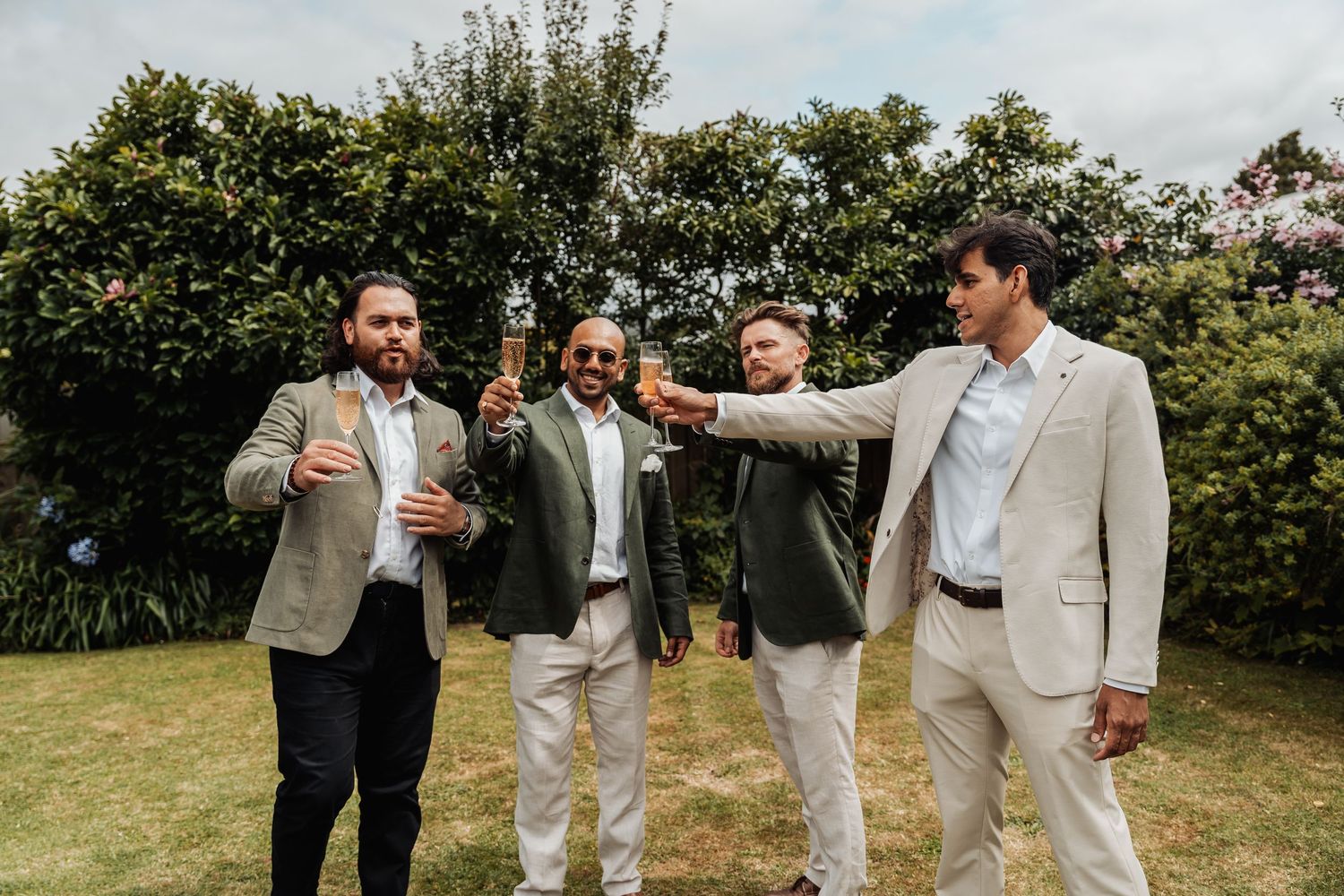 Four groomsmen in suits toast with champagne bottles in a sunny garden before a wedding.