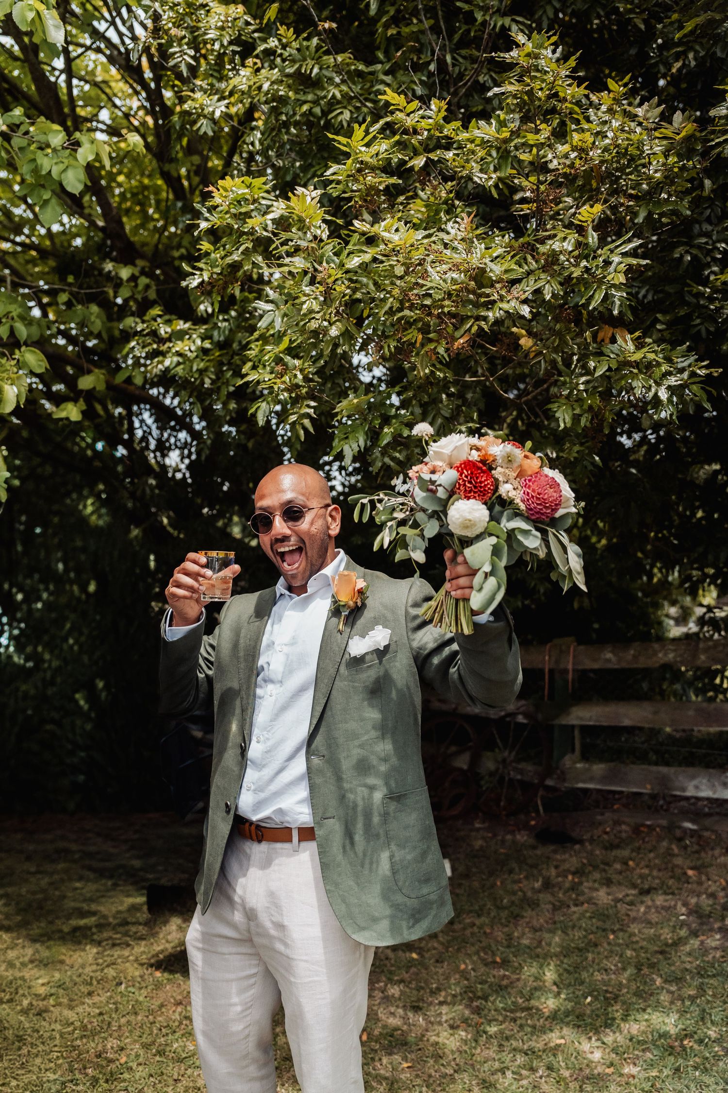 Excited groomsman in green blazer raising drink and bouquet in celebration outdoors.