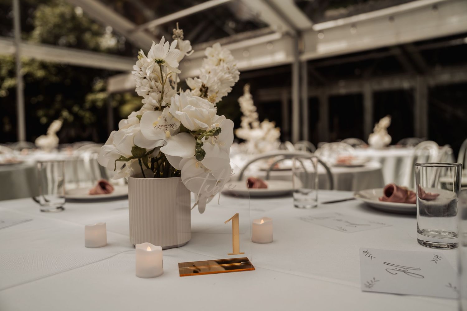 Elegant wedding table centerpiece with white floral arrangement, candles, and gold table number on white linen.