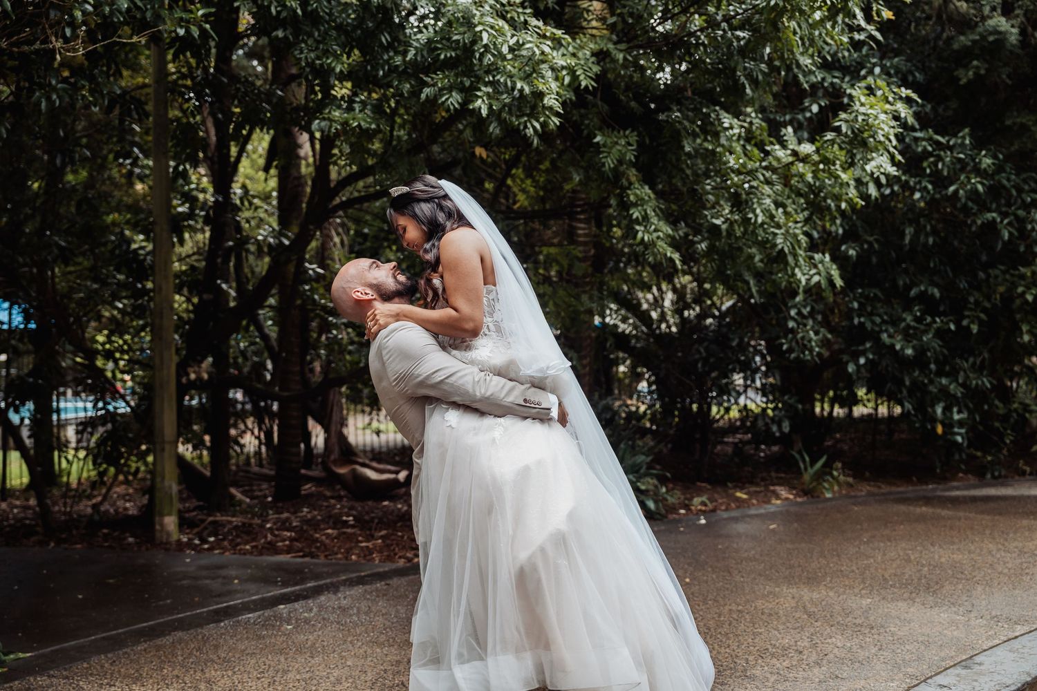 A groom lifts and dips his bride in a white wedding gown and veil among lush green trees outdoors.
