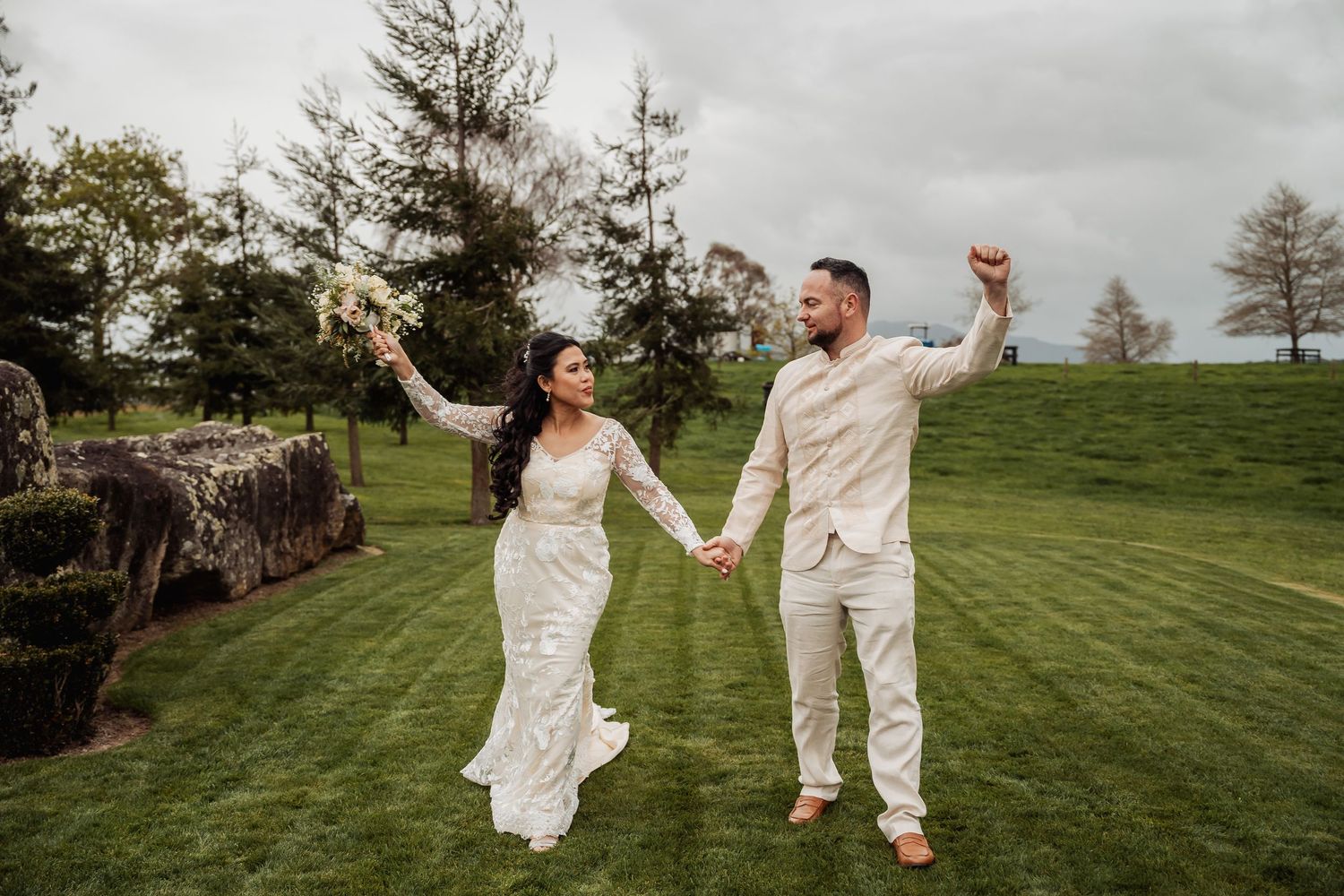 Newlyweds dance and celebrate together on a grassy hillside under cloudy skies.