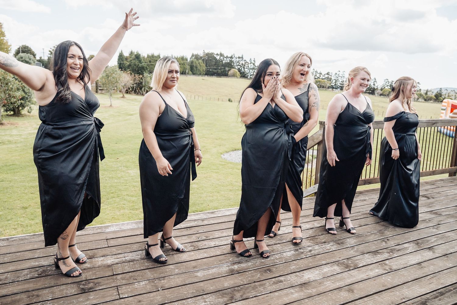 Five women in matching black dresses pose joyfully on a wooden deck overlooking grassy fields.