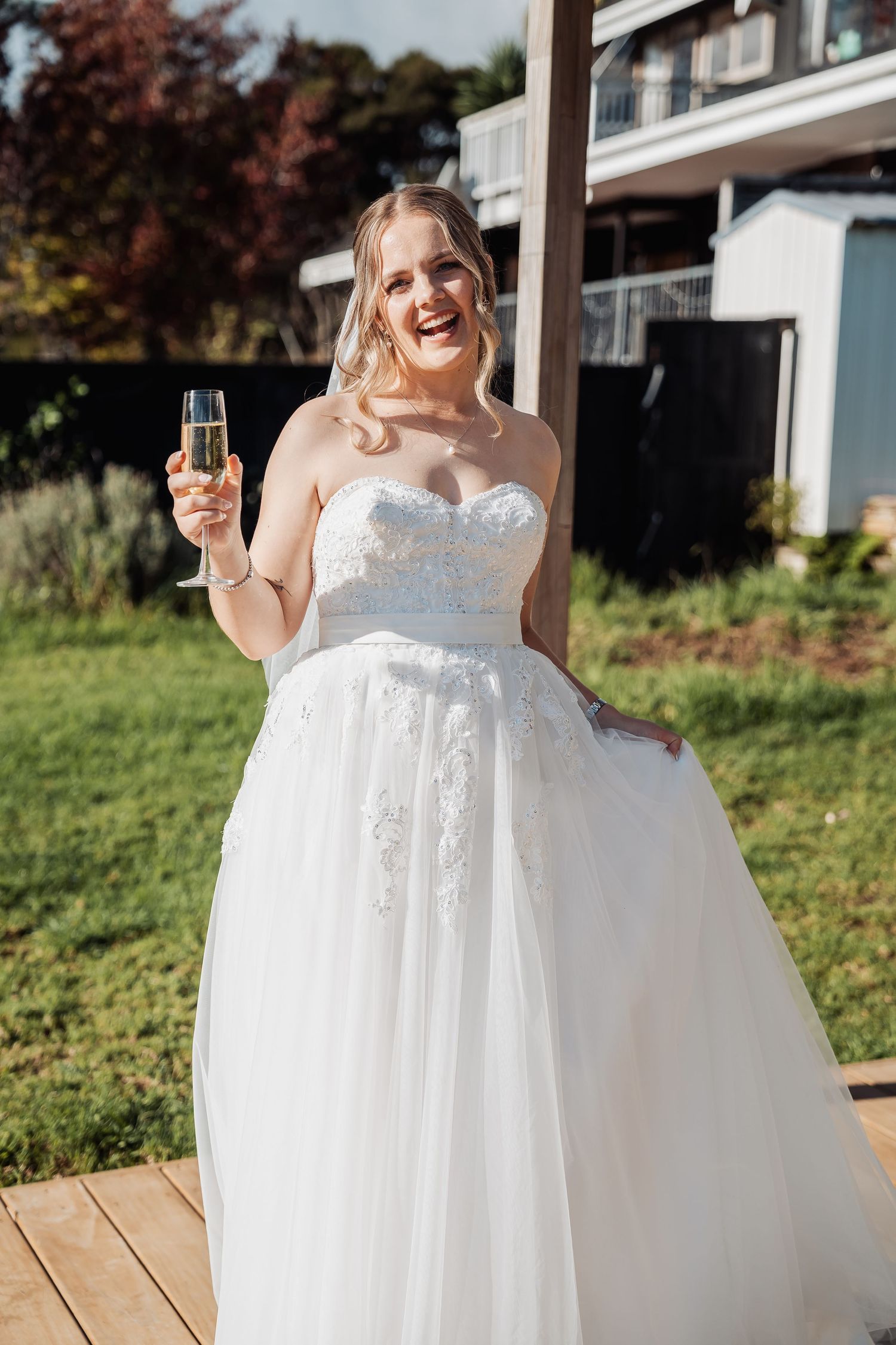 A bride in a strapless lace wedding gown stands on a wooden deck holding a champagne glass and smiling joyfully.