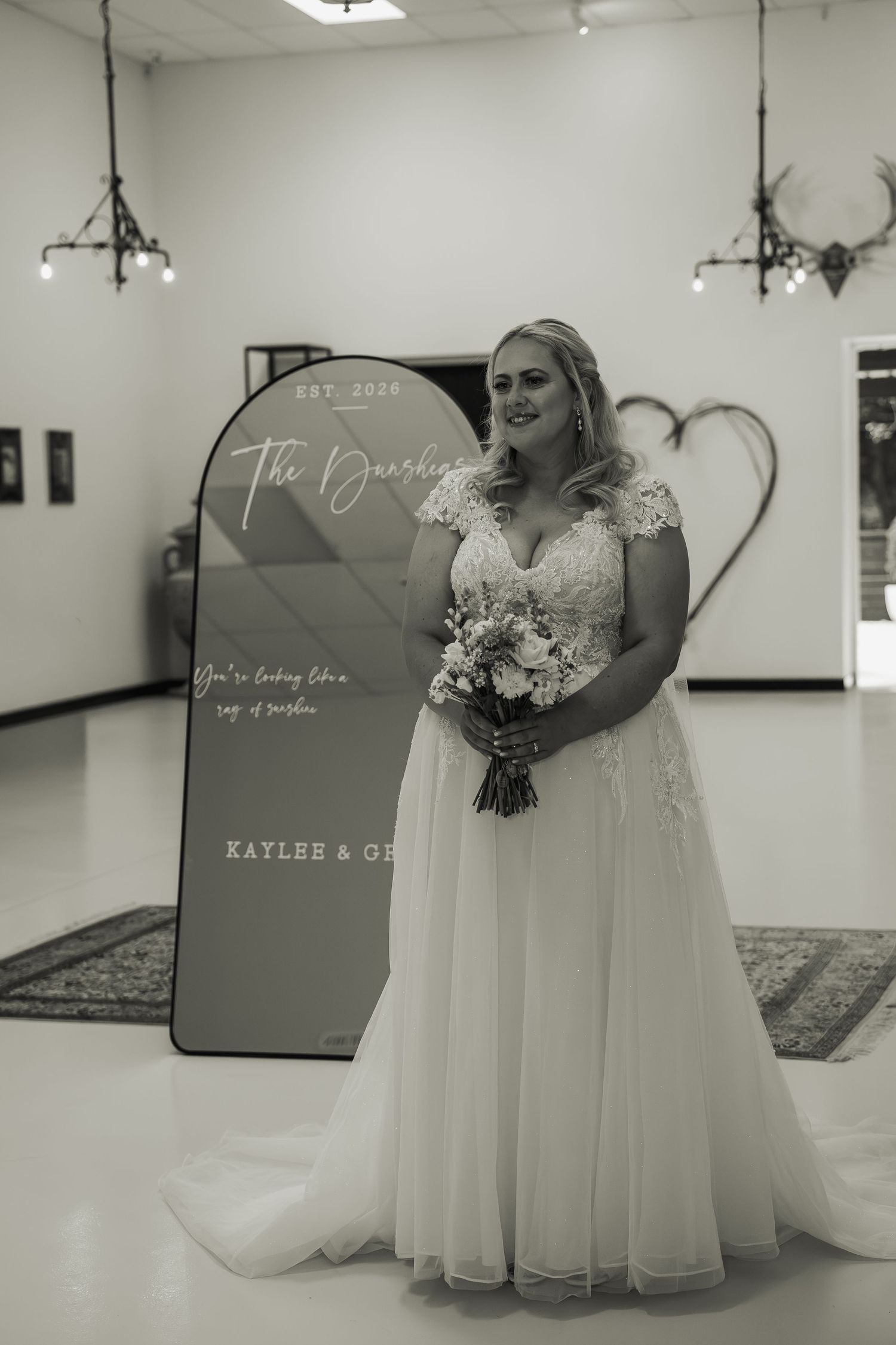Black and white portrait of bride in lace wedding gown holding bouquet beside personalized wedding welcome sign.