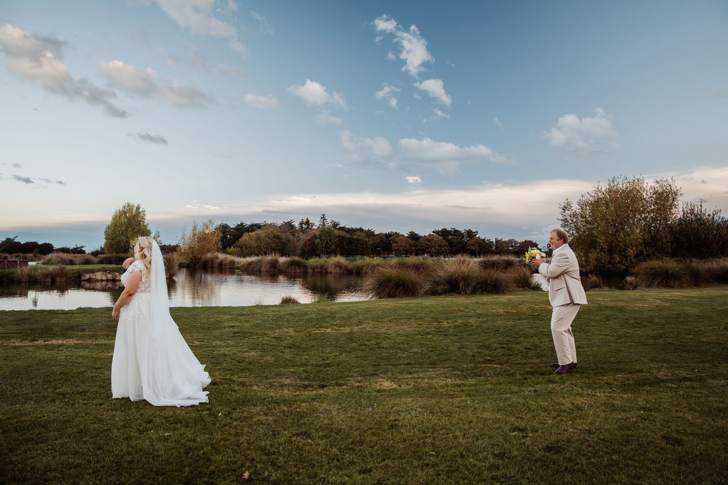 Bride and groom share a joyful moment on green lawn beside tranquil lake at golden hour.