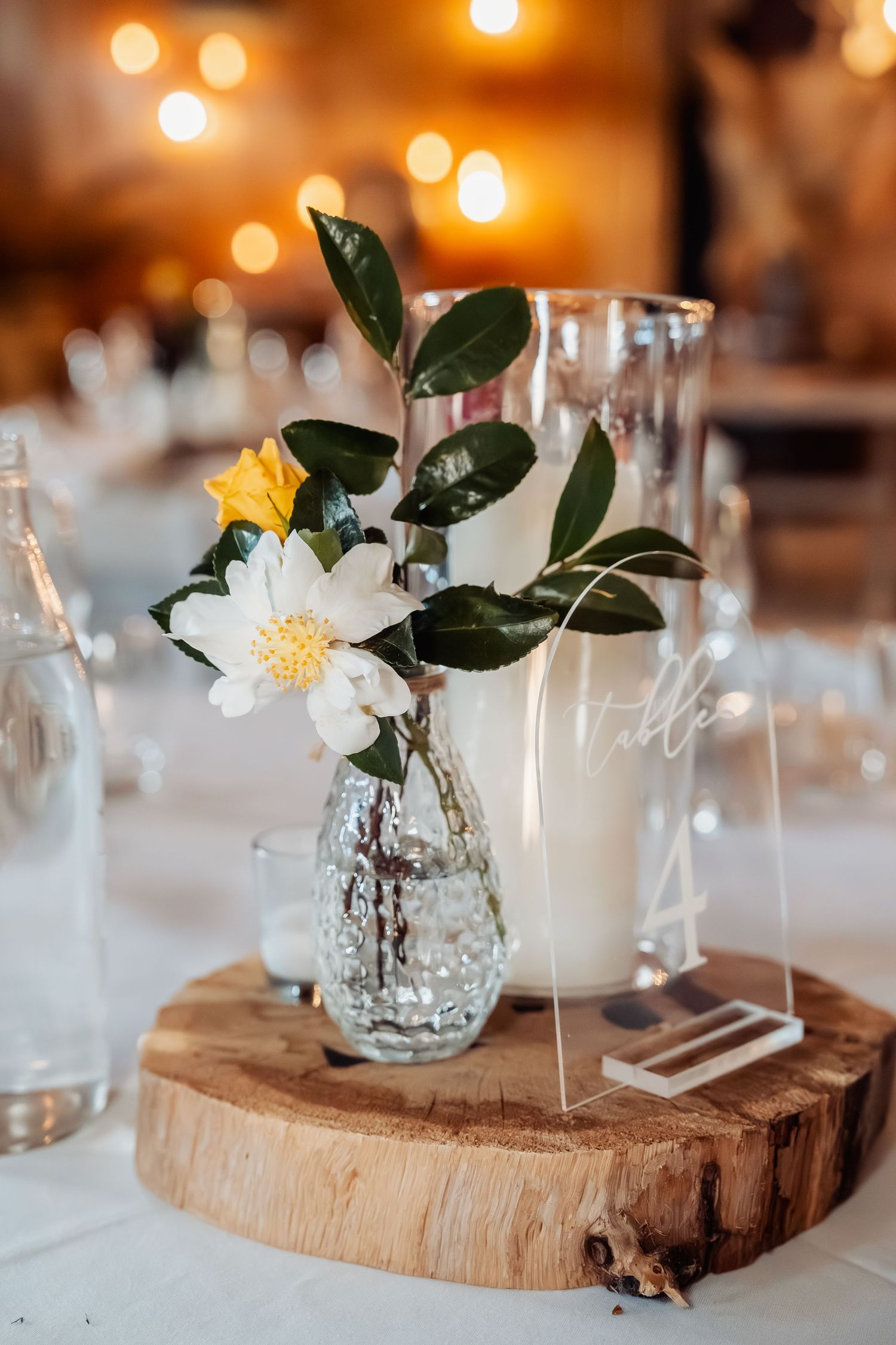 Elegant wedding table centerpiece with white camellia flowers in glass vase on wooden slice with acrylic table number.