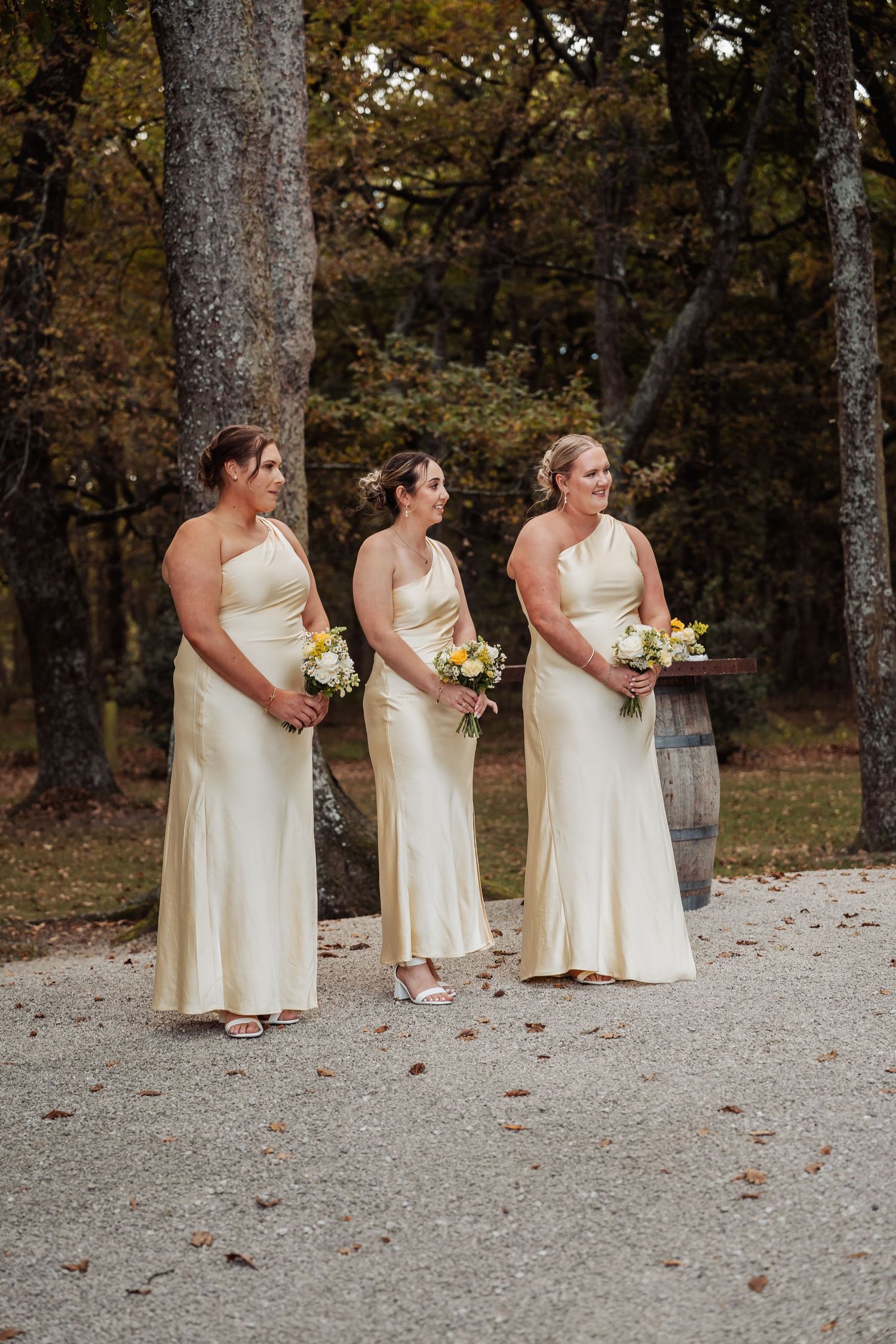 Three bridesmaids in matching cream one-shoulder gowns hold yellow and white bouquets outdoors among autumn trees.