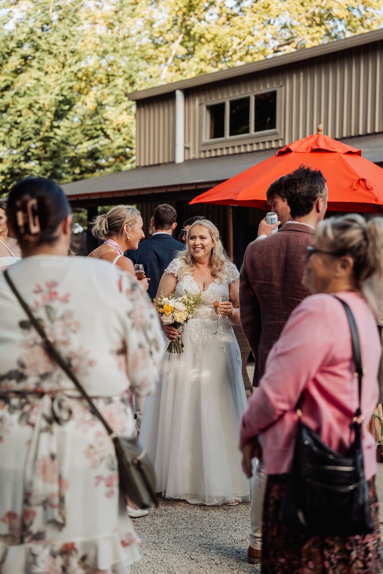 Smiling bride in white gown holding yellow bouquet mingles with wedding guests outdoors near a red umbrella.