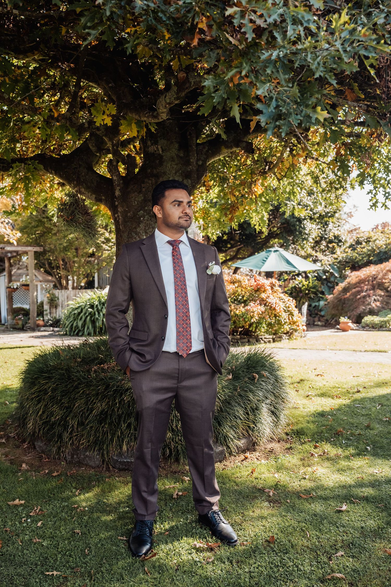 Groom in brown suit with floral tie stands confidently under autumn tree in lush garden setting.