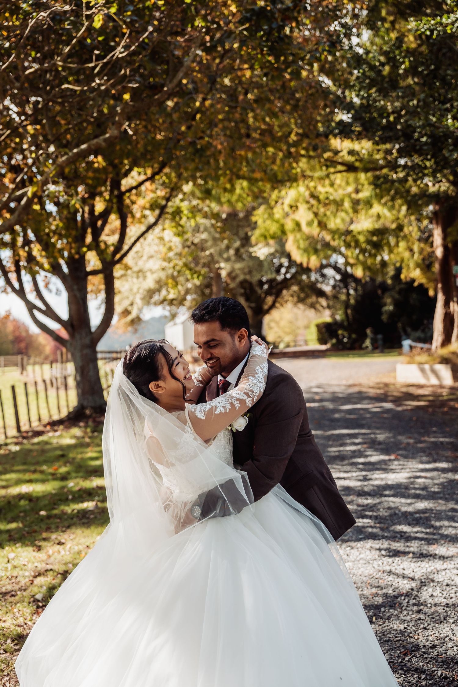 Bride and groom share a joyful embrace on a sunlit tree-lined path in stunning autumn wedding attire.